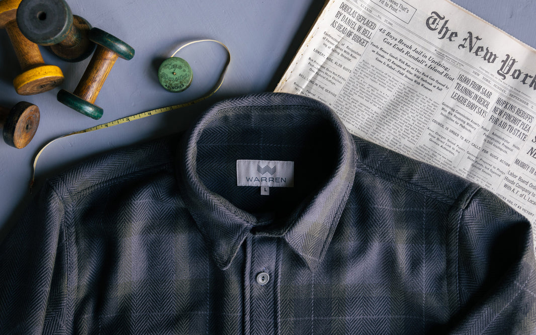 A blue flannel Warren shirt sitting on top of an old newspaper and vintage measuring tape.