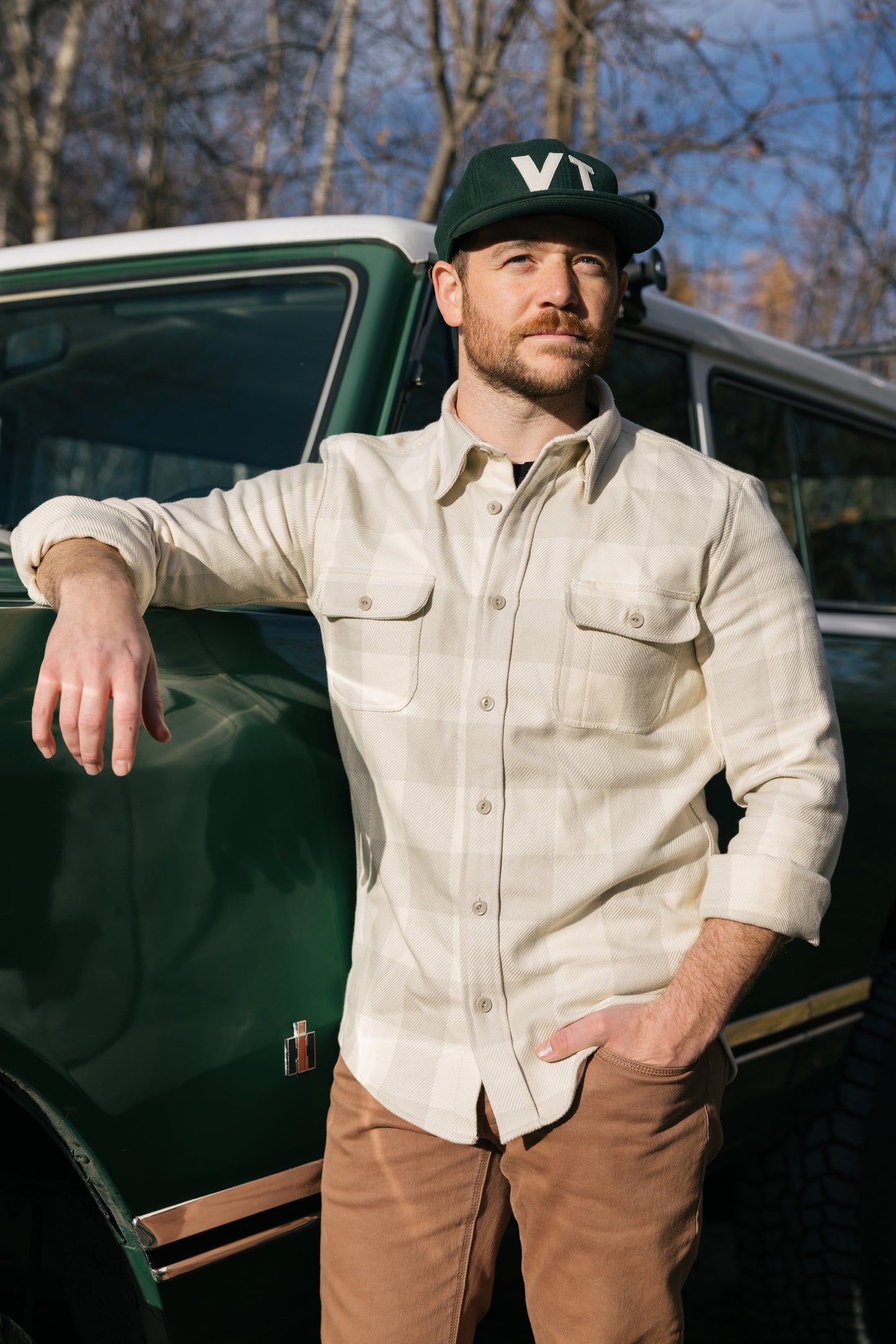 Man leaning against a truck wearing a tan plaid Six Season flannel shirt and Farm League hat by Warren.