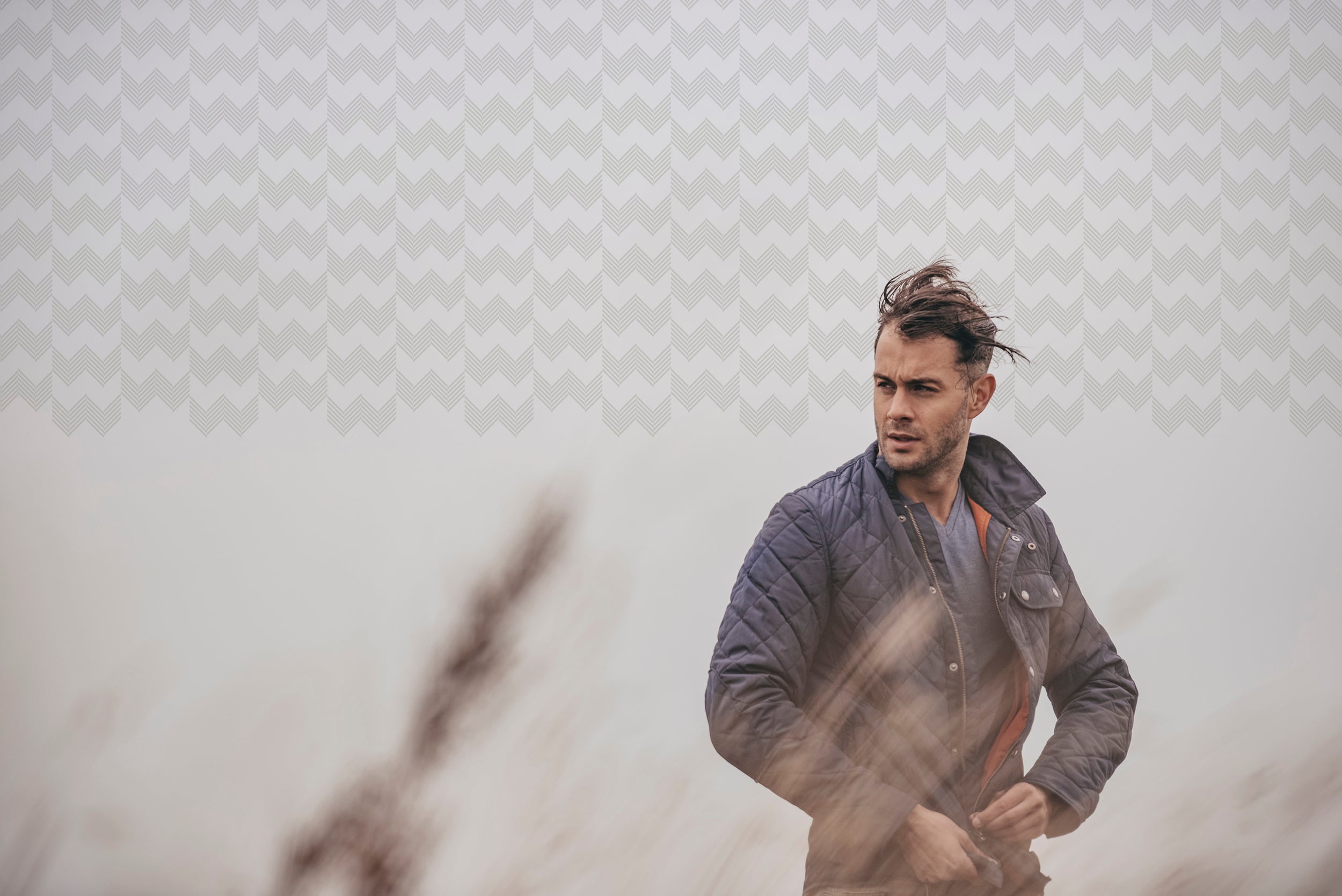 Man zipping up a blue jacket standing in tall grass wind blown hair